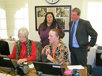 Four people in photo. Two women sitting at computer and two people (one woman and one man) looking on.