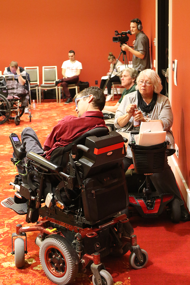 Group of people with various disabilities listening to the panel. A person holding a camera is also present in the photo.