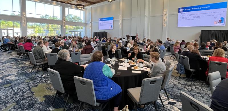 Large conference room filled with people seated at round tables during a group training or workshop, with presentation slides displayed on large screens.