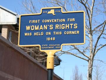 Photo of historical sign (navy with gold writing). Source NPS. Text reads: "New York First Convention for Women's Rights was held on this corner 1848, State Education Department 1932"